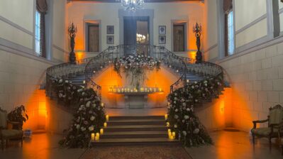 A grand double staircase with ornate railings is decorated with flower arrangements and candles. Warm lighting highlights the steps and walls, while our DJ services Suffolk and Nassau County add to the elegant hall’s romantic, inviting atmosphere.