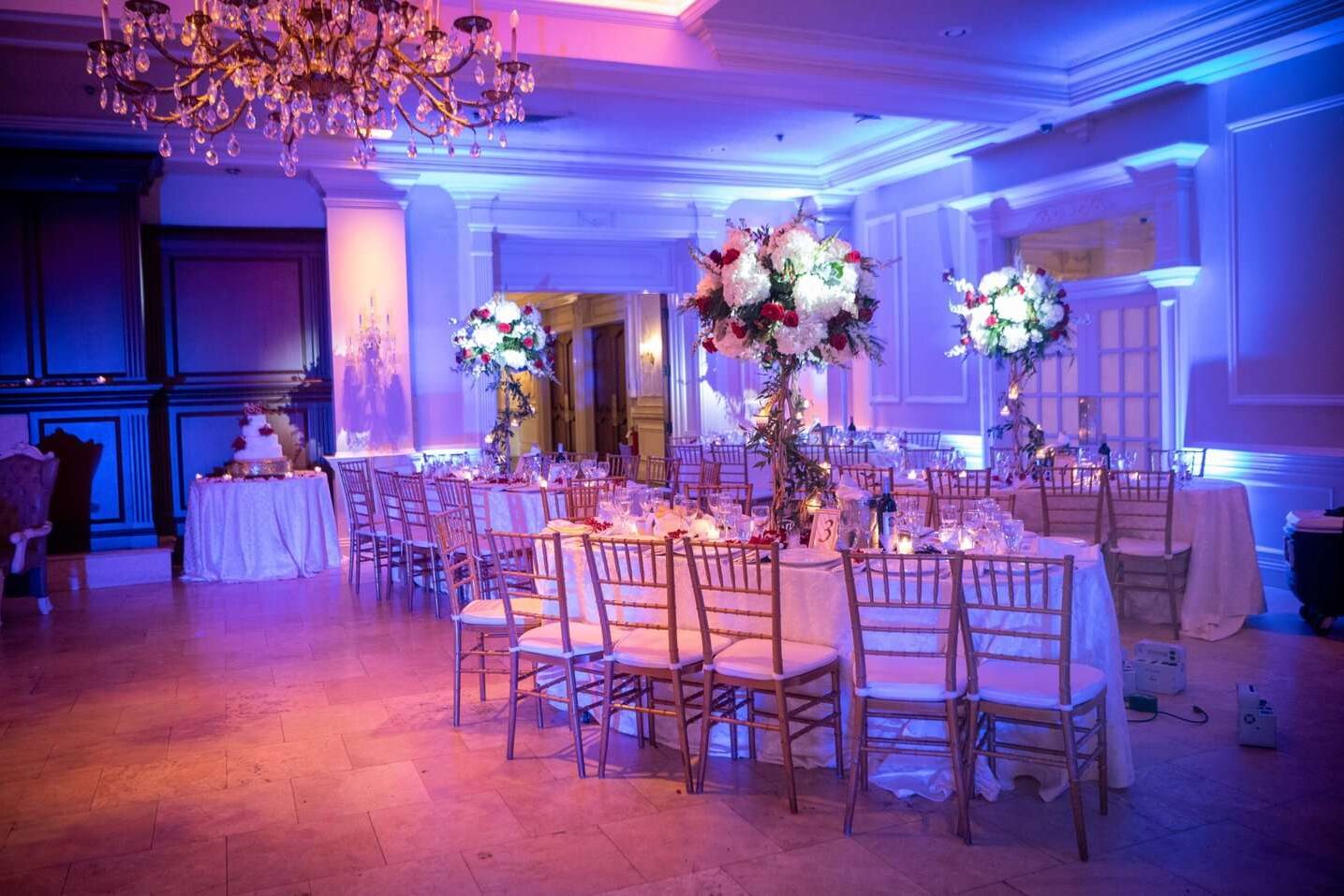 Elegant banquet hall set for an event in NY, with long tables, gold chairs, tall floral centerpieces, glowing purple-blue lighting, and a chandelier overhead. A decorated cake is visible—perfect for dj services Suffolk and Nassau County.
