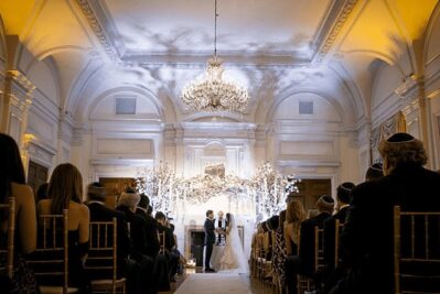 A couple stands under a decorated chuppah at the front of an elegant, chandelier-lit room in NY, surrounded by guests at a wedding ceremony—perfect for those seeking entertainment services in Suffolk and Nassau County, NY.