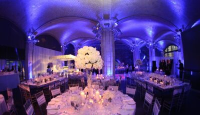 A large, elegant event hall with arched ceilings lit in blue and purple hues, round tables set with white flowers and candles, and silver chairs arranged for a formal gathering or wedding reception with expert DJ services Suffolk and Nassau County.