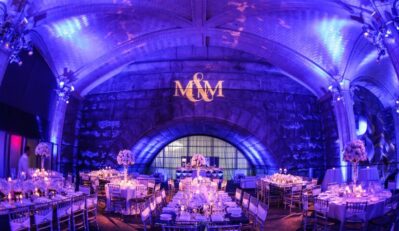 A lavishly decorated NY event space with purple lighting, elegant dinnerware, floral centerpieces on round and rectangular tables, and “M&M” projected on a stone wall—perfect for celebrations with DJ services in Suffolk and Nassau County, NY.