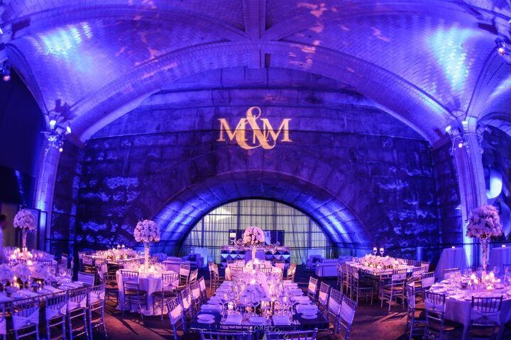 A lavishly decorated NY event space with purple lighting, elegant dinnerware, floral centerpieces on round and rectangular tables, and “M&M” projected on a stone wall—perfect for celebrations with DJ services in Suffolk and Nassau County, NY.