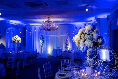 Elegant banquet hall decorated for a wedding reception with blue lighting, round tables set with flowers and candles, tall floral centerpieces, silver chairs, a chandelier overhead, and top entertainment services Suffolk and Nassau County.
