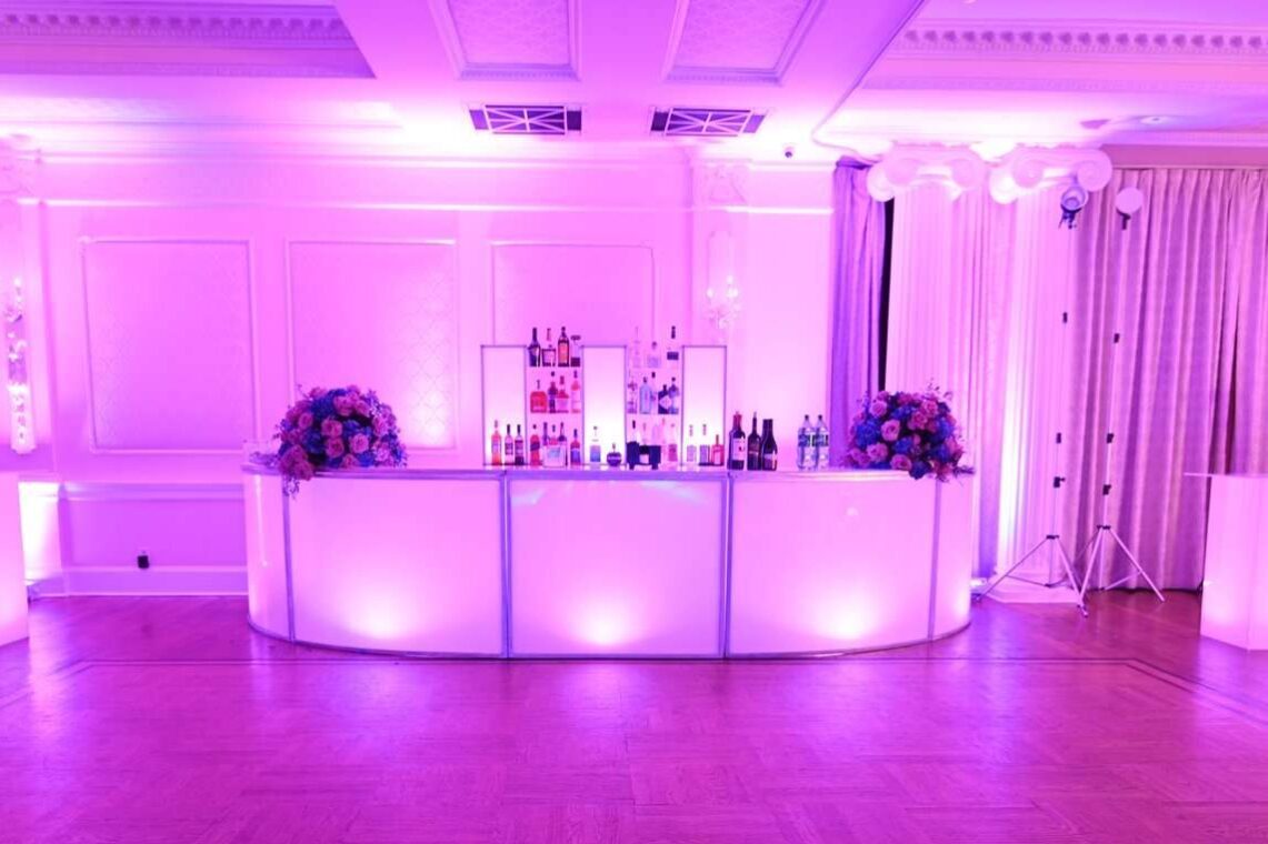 A modern bar with illuminated pink and purple lighting, floral arrangements, and bottle-lined shelves in a spacious room with white walls and wood flooring—perfect for entertainment services Suffolk and Nassau County, NY.