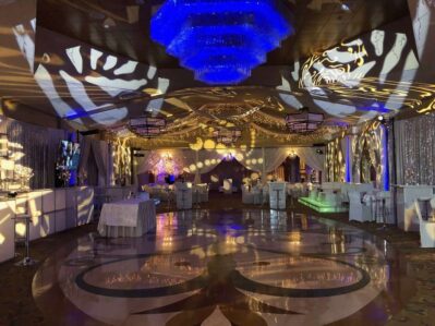 A lavish event hall in NY, decorated with shimmering lights, draped white fabric, and patterned spotlight projections. Enhance your celebration with top entertainment services Suffolk and Nassau County as guests mingle among elegant tables and lounge seating.