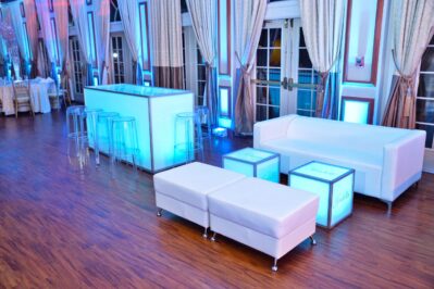 Modern lounge area with white sofas, ottomans, and cube tables, accented by blue LED lighting. Clear bar stools surround a glowing bar on a wood floor—perfect for NY,NY entertainment services in Suffolk and Nassau County.