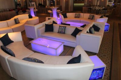 Curved white sofas with dark throw pillows are arranged around glowing purple tables in a modern NY lounge, perfect for entertainment services Suffolk and Nassau County, all set on stylish patterned carpet.