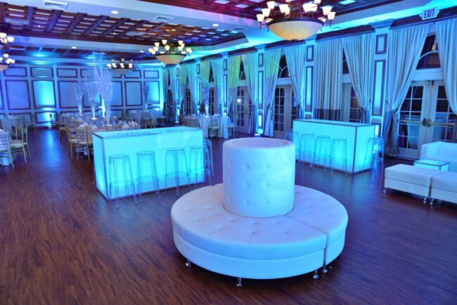 Elegant event hall in NY with modern blue lighting, white tufted seating, wood flooring, tall windows, and chandeliers overhead. Perfect for special occasions, with dj services Suffolk and Nassau County, NY available for an unforgettable celebration.