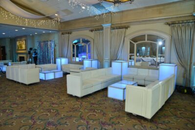 A spacious event hall with elegant decor, featuring white leather sofas and glowing cube tables on a patterned carpet—perfect for entertainment services Suffolk and Nassau County, NY—with draped curtains and large mirrors enhancing the ambiance.
