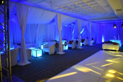 A modern lounge area with white chairs and draped fabric, blue accent lighting, cube-shaped illuminated tables, and decorative lanterns—perfect for NY events or dj services Suffolk and Nassau County—offers an elegant, sophisticated atmosphere.