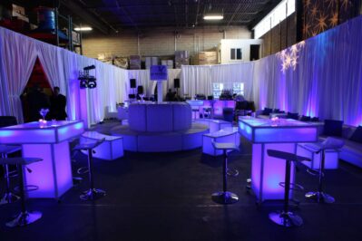 A modern NY event space with white furniture, illuminated tables, and purple-blue ambient lighting. Sheer curtains create a lounge vibe in a large industrial warehouse—perfect for entertainment or DJ services in Suffolk and Nassau County.