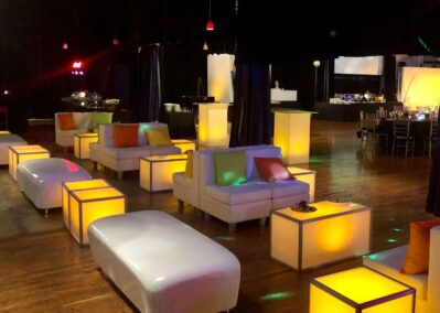 A modern event space with white lounge furniture, colorful throw pillows, and illuminated yellow tables on a wooden floor. Ideal for gatherings, it offers entertainment services in Suffolk and Nassau County, NY. Soft hanging lights complete the festive atmosphere.