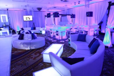 A modern event lounge featuring glowing purple and blue lighting, white curved sofas, illuminated tables, and draped curtains—perfect for upscale gatherings with premium entertainment services in Suffolk and Nassau County, NY.