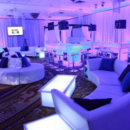 A modern event lounge featuring glowing purple and blue lighting, white curved sofas, illuminated tables, and draped curtains—perfect for upscale gatherings with premium entertainment services in Suffolk and Nassau County, NY.