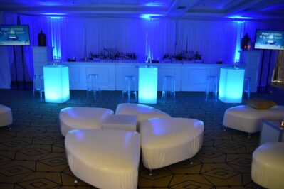 A modern NY lounge with glowing blue-lit tables, clear barstools, and white rounded couches. The softly illuminated space is perfect for entertainment services in Suffolk and Nassau County, NY. The sleek bar shines in the background.