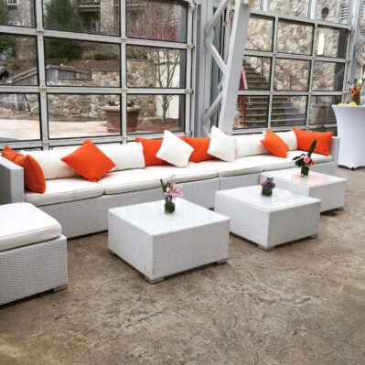 A long white wicker sofa with orange and white throw pillows sits in a bright NY room with glass garage doors. Three matching coffee tables hold small vases with flowers—perfect for entertainment services in Suffolk and Nassau County, NY.