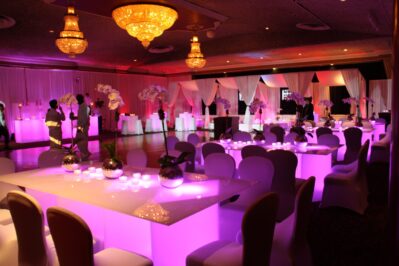 A decorated banquet hall in NY with glowing purple-lit tables, white chairs, elegant centerpieces, and chandeliers. People mingle near draped white fabric and illuminated booths. DJ services Suffolk and Nassau County, NY add to the festive atmosphere.