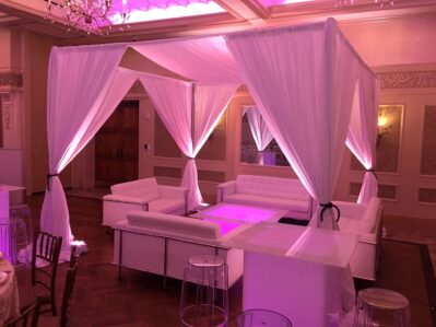 A lounge area with white sofas and clear stools arranged under a canopy of sheer white drapes, softly lit with purple lighting in an elegant indoor venue—perfect for NY events or dj services Suffolk and Nassau County, NY.