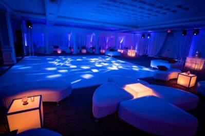 A spacious, modern event hall in NY with white seating, glowing tables, and blue ambient lighting. Decorative light patterns are projected onto the large, empty dance floor—ideal for events featuring top DJ services Suffolk and Nassau County, NY.