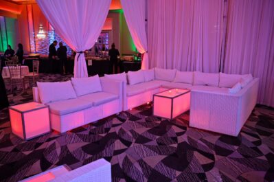 A lounge area with white sofas and tables lit by pink lights, set against white drapery. The space delivers NY,NY style elegance—perfect for entertainment services in Suffolk and Nassau County—with a patterned carpet and a lively bar scene.