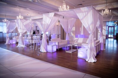 Elegant NY event space with white draped canopies, modern sofas, illuminated cubes, and chandeliers—offering a soft, upscale atmosphere enhanced by natural light, purple accent lighting, and top entertainment services Suffolk and Nassau County.