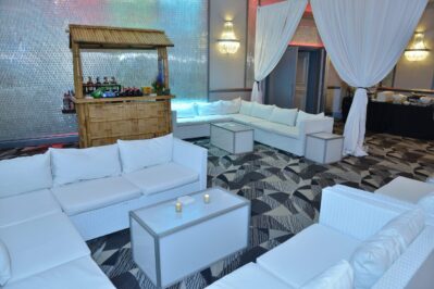 A lounge area with white couches and a small table with candles sits on a patterned carpet. A bamboo bar stands in the background, while draped white curtains and modern chandeliers set the tone—perfect for events with dj services Suffolk and Nassau County, NY.