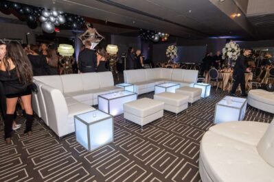 Modern lounge area at an event, featuring white couches and ottomans with illuminated cube tables—perfect for entertainment services Suffolk and Nassau County, NY—surrounded by guests in black and silver decor, balloons, and floral arrangements.