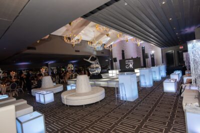 A modern NY,NY event space with white lounge seating, illuminated cube tables, chandeliers, and a mirrored sculpture under dramatic lighting. Ideal for entertainment services in Suffolk and Nassau County. "CLUB CM" stage features in the background.