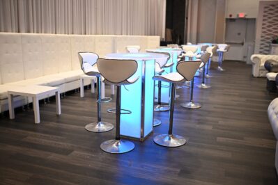 Modern lounge with white high-backed seating, illuminated tall tables, and chrome barstools. Dark wood floors and light curtains create a sleek vibe—perfect for events featuring DJ services in Suffolk and Nassau County, NY.