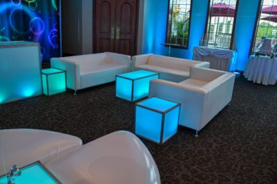 A modern lounge area in NY with white sofas and glowing blue square tables, set in a spacious room with large windows, dark carpet, decorative blue lighting, and available dj services Suffolk and Nassau County, NY.