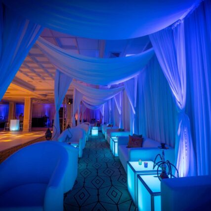 A luxurious lounge area with white sofas, draped white curtains, and glowing blue cube tables is bathed in blue and purple lighting—perfect for entertainment services Suffolk and Nassau County seeking an elegant, intimate atmosphere.