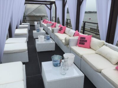 A lounge area with white wicker sofas and ottomans, decorated with pink "NYC PRIDE 2015" pillows, set under a canopy with white curtains by the NY waterfront. Perfect for events, especially with DJ services Suffolk and Nassau County can provide.