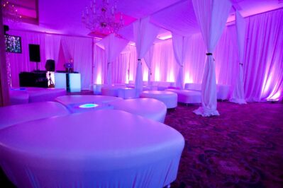 A modern lounge with white cushioned seating, draped white fabric, and purple mood lighting. Chandeliers hang from the ceiling, and a DJ booth offers top-tier dj services Suffolk and Nassau County. The atmosphere is elegant and upscale.