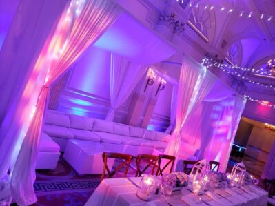 A luxurious NY event space decorated with white drapes, glowing purple and pink ambient lighting, a long table set with flowers and candles, a white lounge area under chandeliers, and premium entertainment services Suffolk and Nassau County.