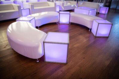 Curved white couches and glowing cube-shaped tables arranged on a wooden floor in a modern, stylish lounge setting. Perfect for NY,NY entertainment services in Suffolk and Nassau County, the cubes emit soft purple light for a contemporary ambiance.