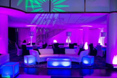 A modern lounge with white furniture, glowing tables, and purple and blue ambient lighting. Stairs are visible in the background, and lamps with shades add to the decorative atmosphere—ideal for DJ services in Suffolk and Nassau County, NY.