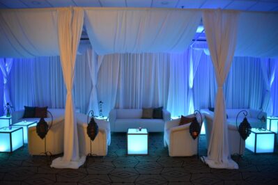 A modern lounge area with white sofas, glowing cube tables, elegant draped curtains, and soft blue lighting—perfect for events featuring top DJ services Suffolk and Nassau County, NY, creating a serene and upscale atmosphere.