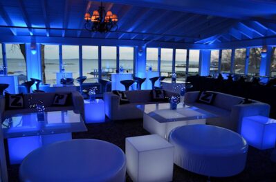 Modern lounge in NY with blue ambient lighting, white cube tables, round ottomans, sofas with lettered pillows, and large windows overlooking a waterfront at dusk—perfect for events featuring top DJ services in Suffolk and Nassau County, NY.