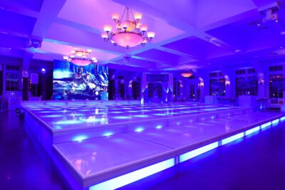 A modern indoor event space in NY features a large, illuminated dance floor glowing with blue and purple lights, elegant chandeliers overhead, and a digital screen displaying a vibrant scene—perfect for entertainment services in Suffolk and Nassau County, NY.