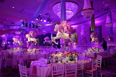 A lavish NY ballroom decorated in purple lighting features round tables with colorful floral centerpieces, elegant chandeliers, and white carousel horse statues adorned with flowers suspended above the tables; perfect for events needing dj services Suffolk and Nassau County,NY.