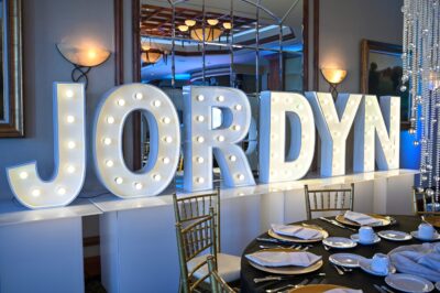Large marquee letters spelling "JORDYN" are lit up behind a table set with plates, cups, and gold chairs. The room, perfect for NY,NY events and entertainment services Suffolk and Nassau County, features mirrors, paintings, and hanging crystal strands.