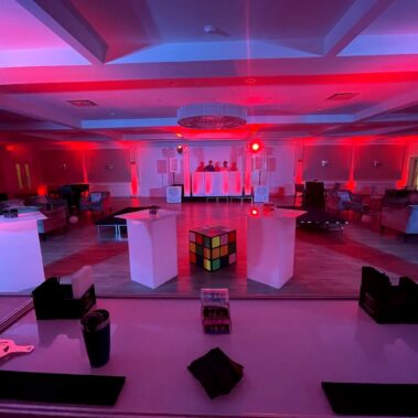 A large event room in NY with red lighting features a DJ booth in the back, a giant Rubik’s cube centerpiece, white pedestal tables, and seating areas—ideal for entertainment services in Suffolk and Nassau County.