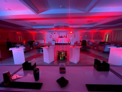 A large event room in NY with red lighting features a DJ booth in the back, a giant Rubik’s cube centerpiece, white pedestal tables, and seating areas—ideal for entertainment services in Suffolk and Nassau County.