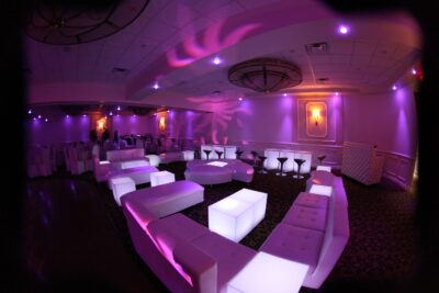 A modern event space with glowing white lounge furniture, illuminated in purple lighting. Ideal for entertainment services Suffolk and Nassau County, NY, the room features round tables, elegant wall sconces, and a stylish patterned carpet.