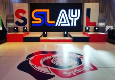 A stage with a large screen displaying "SLAY" in bold letters, surrounded by red, white, and blue decor. The floor features a stylized rose and the word "Sydney," perfect for NY events or DJ services in Suffolk and Nassau County, NY.