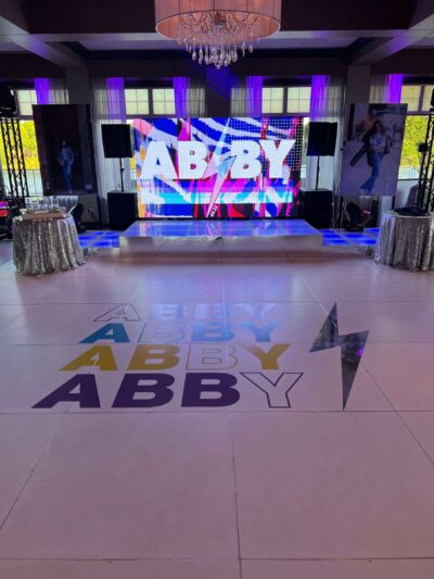 A decorated event space features a white dance floor with colorful "ABBY" text and a lightning bolt, silver-covered tables, and a vibrant LED screen—perfect for dj services Suffolk and Nassau County or entertainment services Suffolk and Nassau County, NY.
