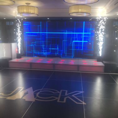 A modern event stage in NY with a large digital screen displaying blue geometric lines, pyrotechnic effects on either side, and a black dance floor with "JACK" in large letters—ideal for dj services Suffolk and Nassau County, NY.