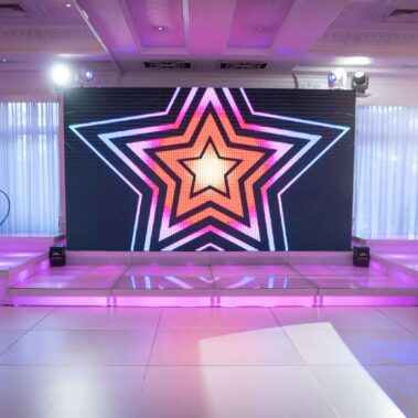 A modern NY event stage with a large LED screen displaying a colorful, layered star pattern. Chandeliers, pink-purple lighting, speakers, and spotlights set the mood—perfect for entertainment services Suffolk and Nassau County.