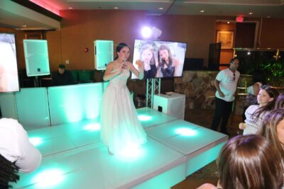 A young woman in a white dress stands on a glowing stage with a microphone, performing at a party with DJ services Suffolk and Nassau County, NY. People watch from the dance floor as a large screen behind her shows two smiling girls.