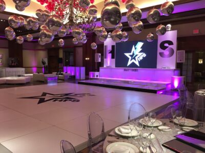 A modern event space with a large dance floor, star and lightning bolt logo, purple lighting, mirrored ball decor, a glittery dining table, and a stage with a big screen—perfect for entertainment services in Suffolk and Nassau County.